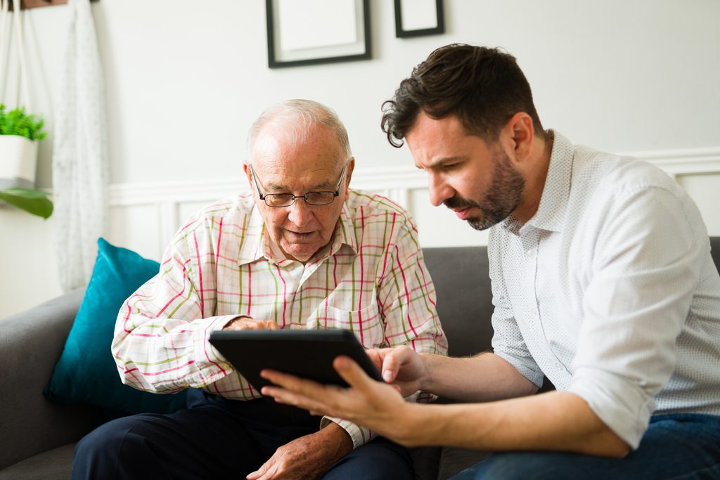 An older man and a younger man looking at a tablet together on a sofa.