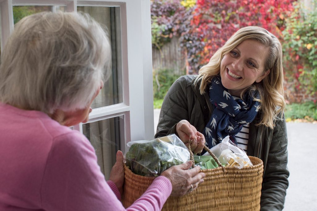 Someone handing a basket of groceries to an older woman at her front door.