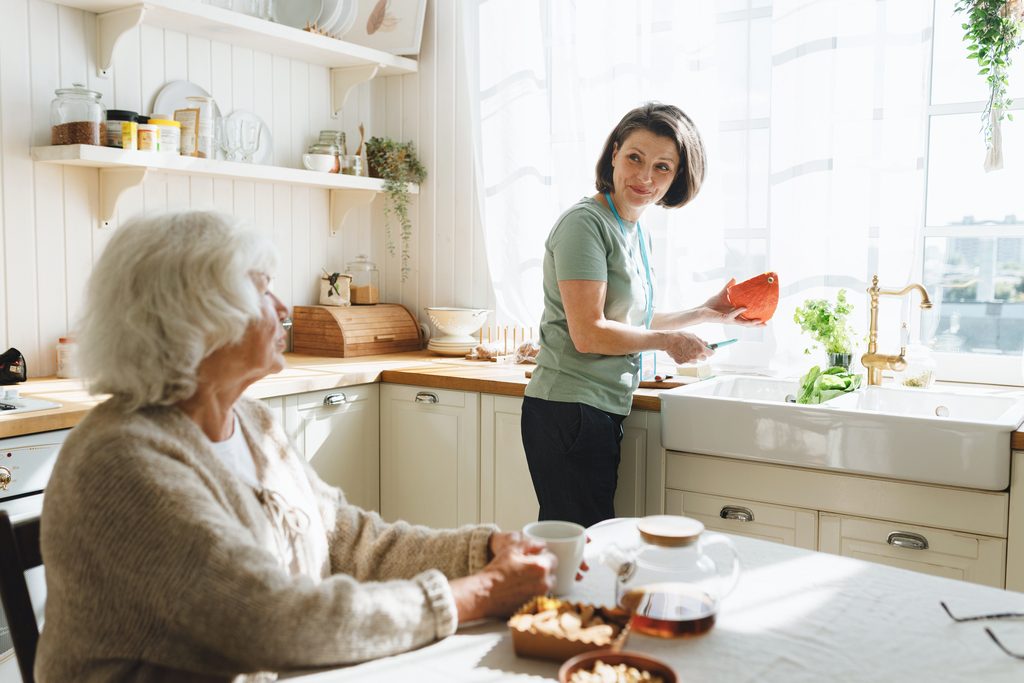 An older woman at her kitchen table while a younger woman washes up at the sink.