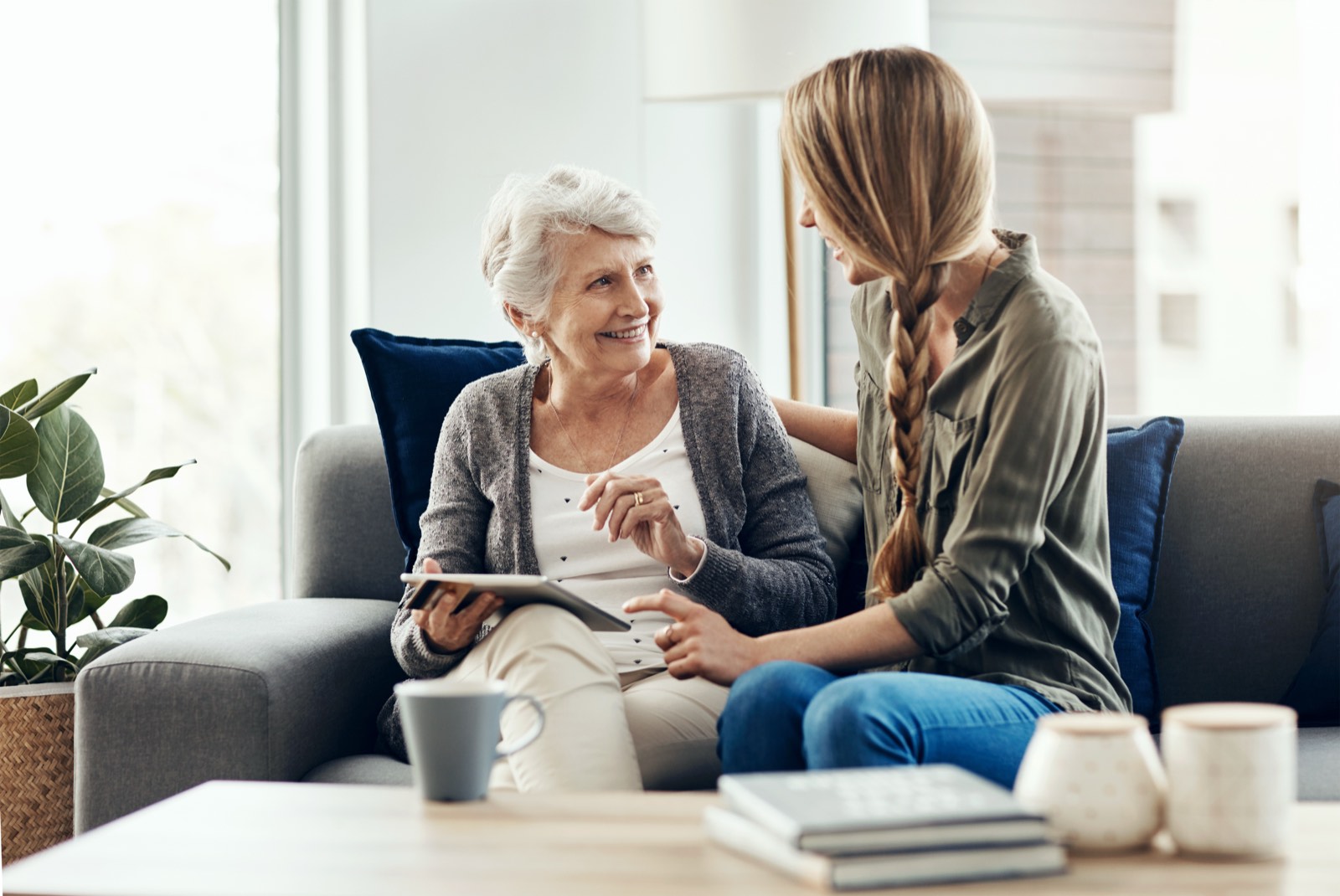 An older woman and a younger woman sitting together on a sofa at home, talking and looking at a tablet.